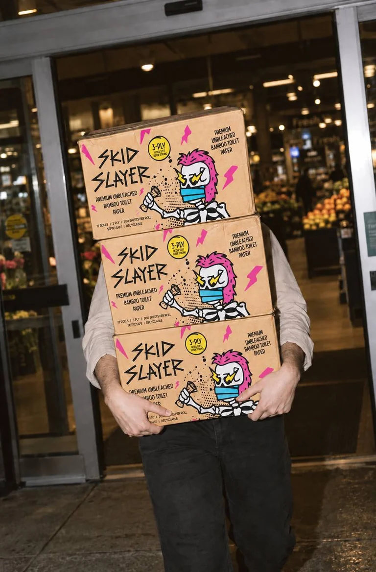 Person carrying three Skid Slayer cardboard boxes, each labelled 12 rolls